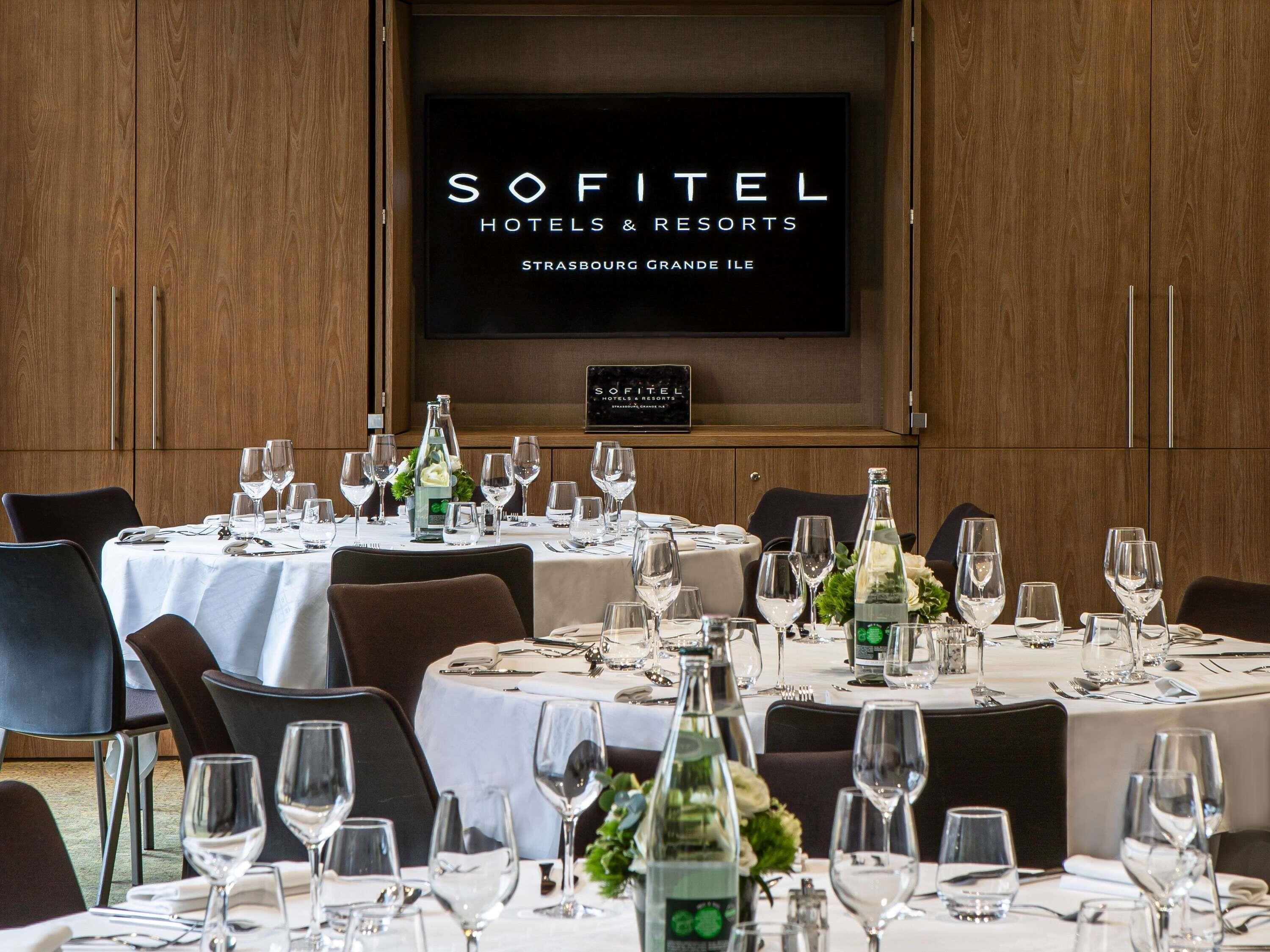 A dining area with tables set for a meal, a television displaying 'SOFITEL HOTELS & RESORTS STRASBOURG GRANDE ILE', and wooden paneling on the walls.