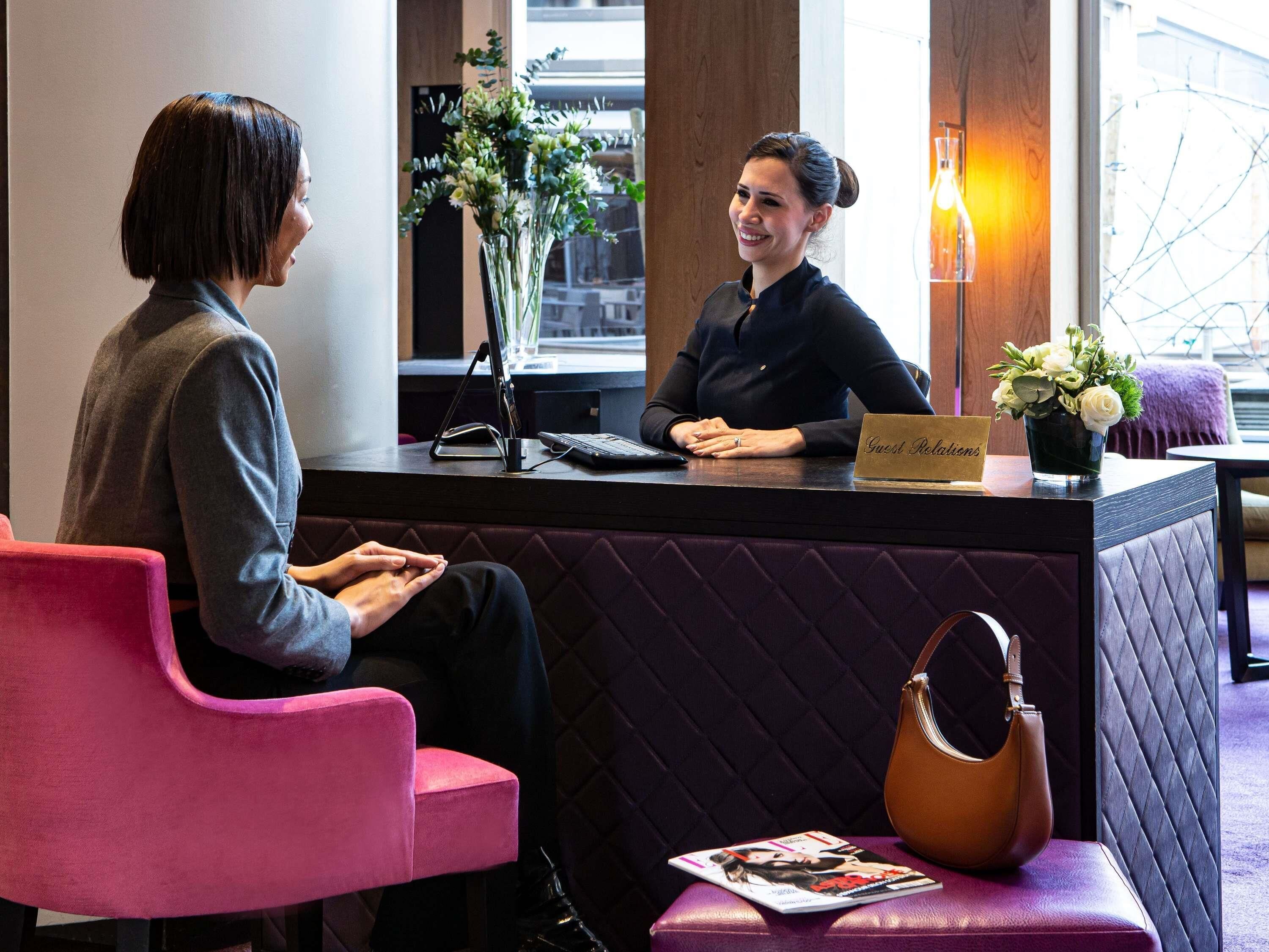 A reception desk with a sign reading 'Guest Relations,' a laptop, a vase with flowers, and a magazine on a bench.