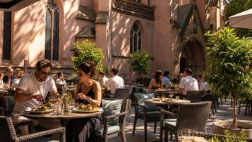An outdoor dining area with wicker chairs and tables, surrounded by buildings with arched windows and a stone facade.