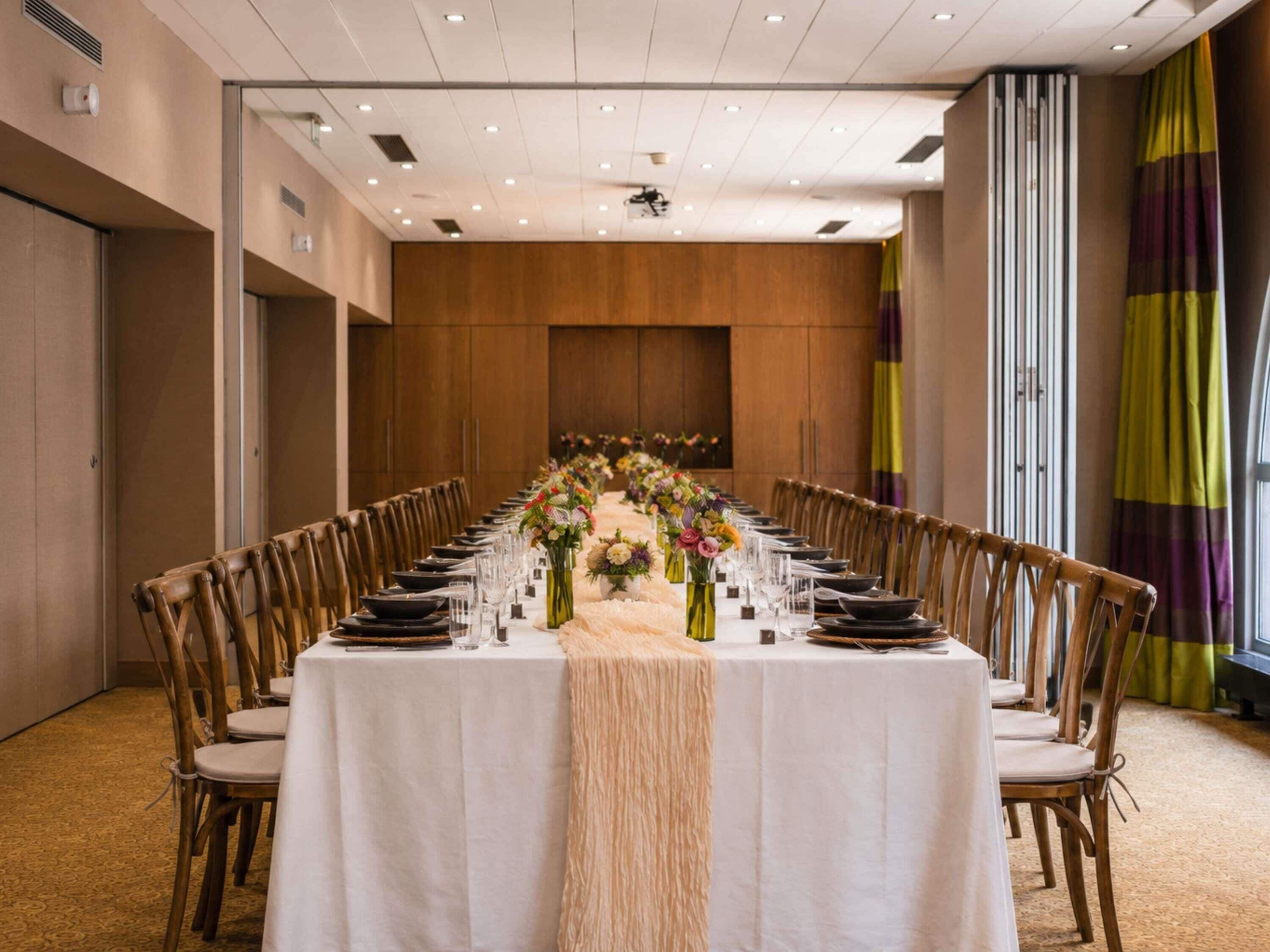 A long conference table set with chairs, floral centerpieces, and a white tablecloth in a room with wooden paneling and a high ceiling.