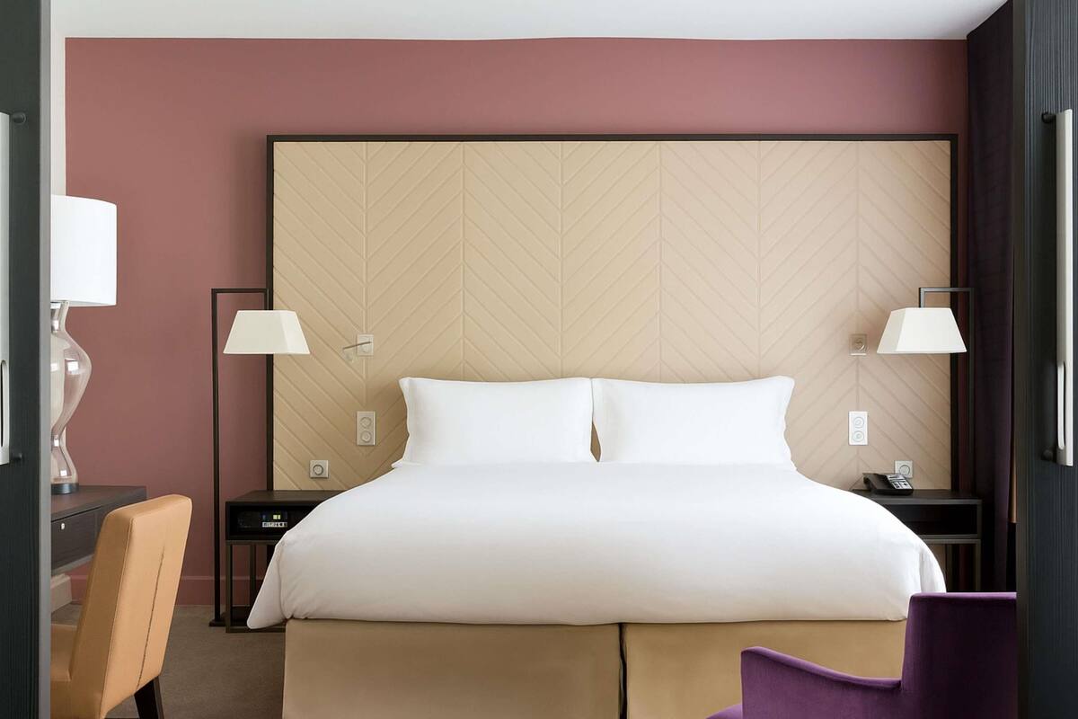 Habitación de hotel con una cama grande, dos mesitas de noche, un escritorio con silla y un sillón púrpura.