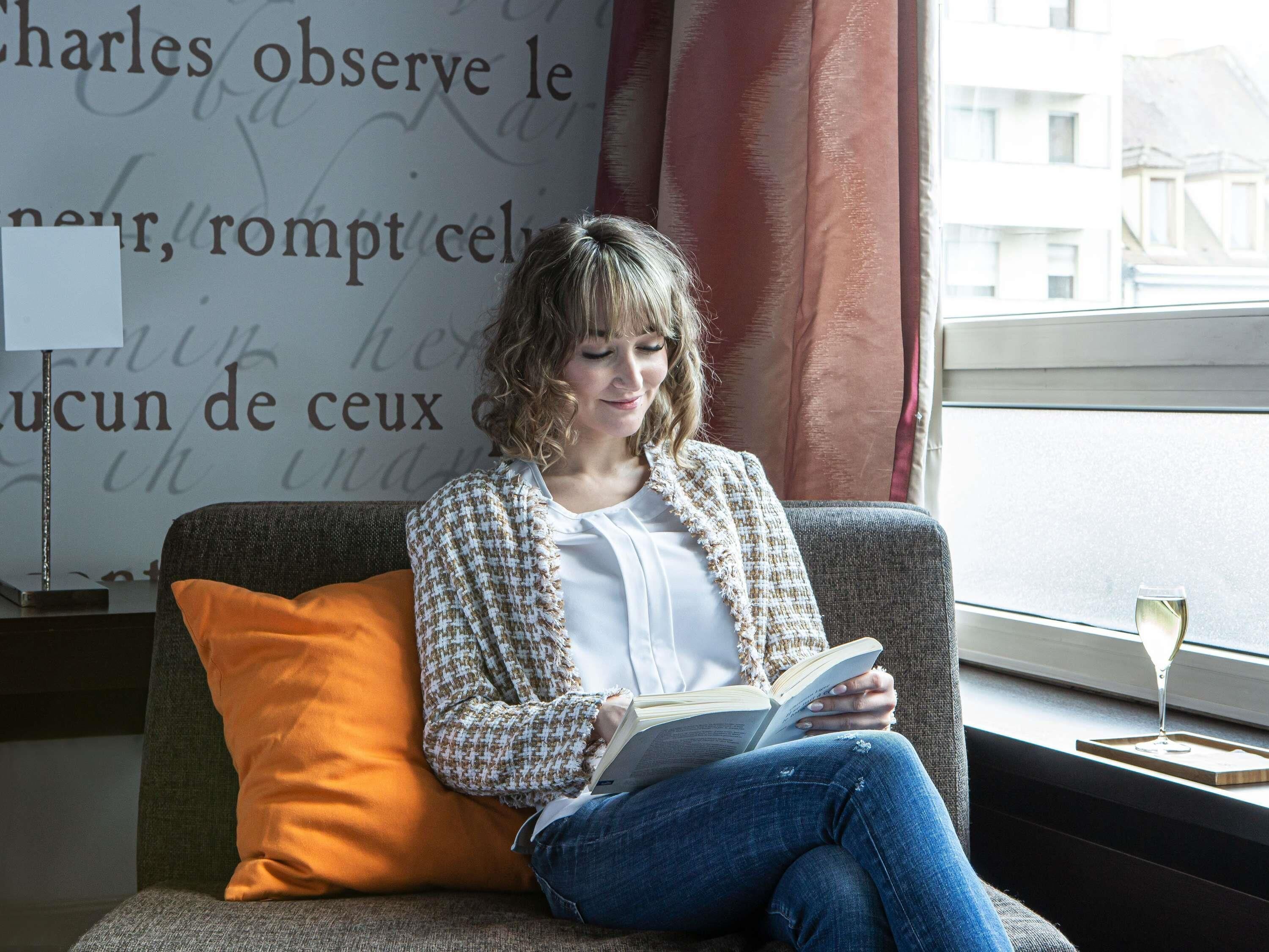A person sitting on a grey sofa with an orange pillow, reading a book, in a room with a window and a lamp.