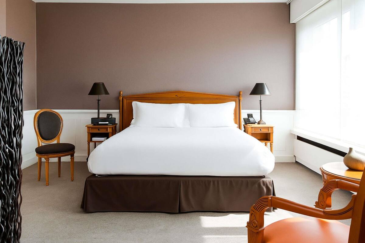 Una habitación de hotel con una cama de madera, dos mesitas de noche, una silla y un ventanal.