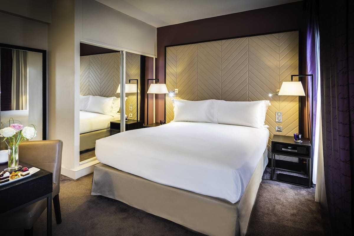 Habitación de hotel con una cama grande, mesitas de noche con lámparas, un espejo, un escritorio con un florero y suelo cubierto de alfombra.