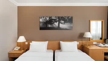 A hotel room with two beds, a nightstand with a lamp, a bedside table with a lamp, a mirror, and a painting of trees on the wall.