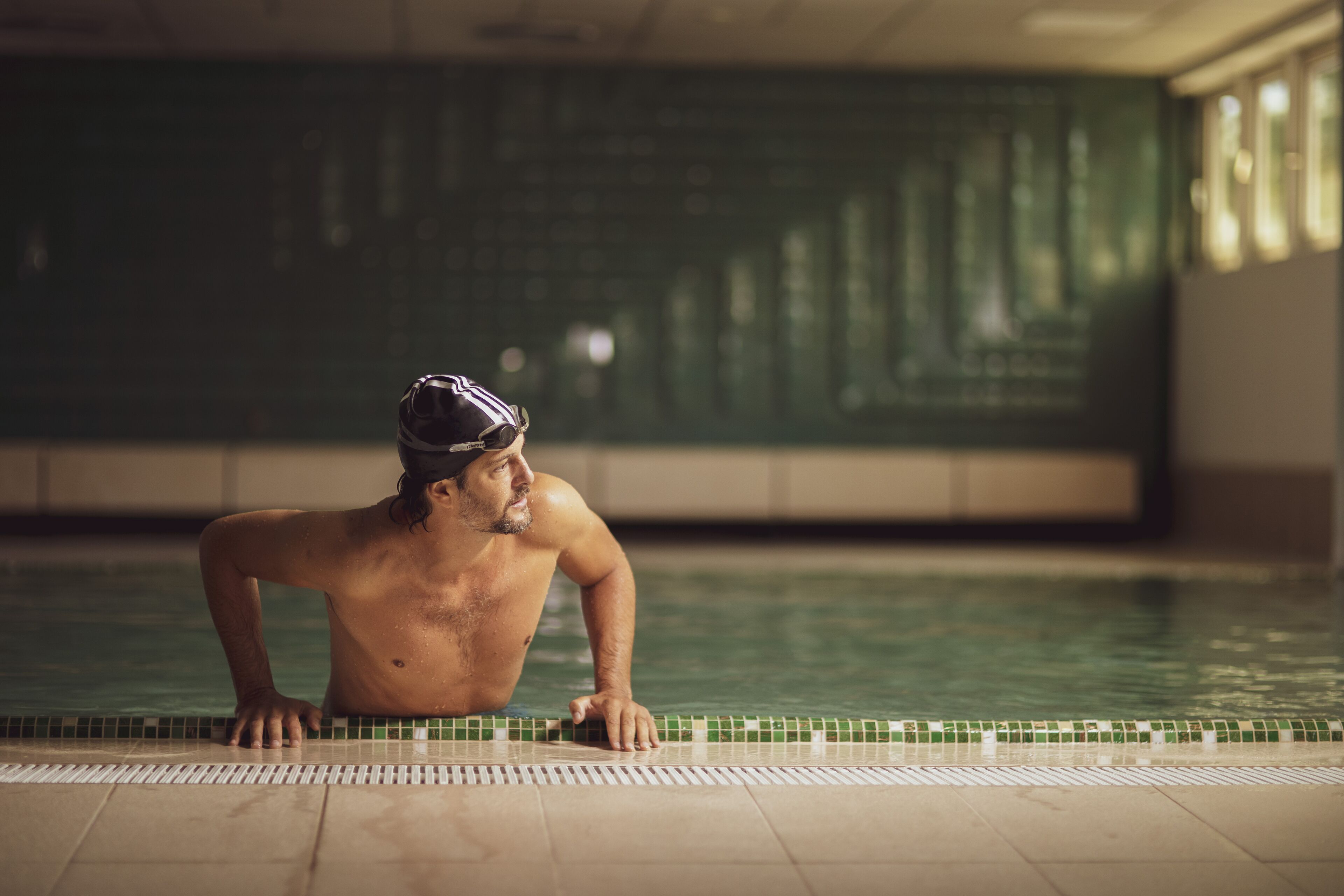 A man in a swimming cap at the edge of a pool with tiled walls and windows in the background.