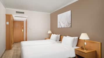 Habitació d'hotel amb dos llits, una taula de nit amb làmpada, un armari de fusta, una ventilació al sostre i una obra d'art penjada a la paret.