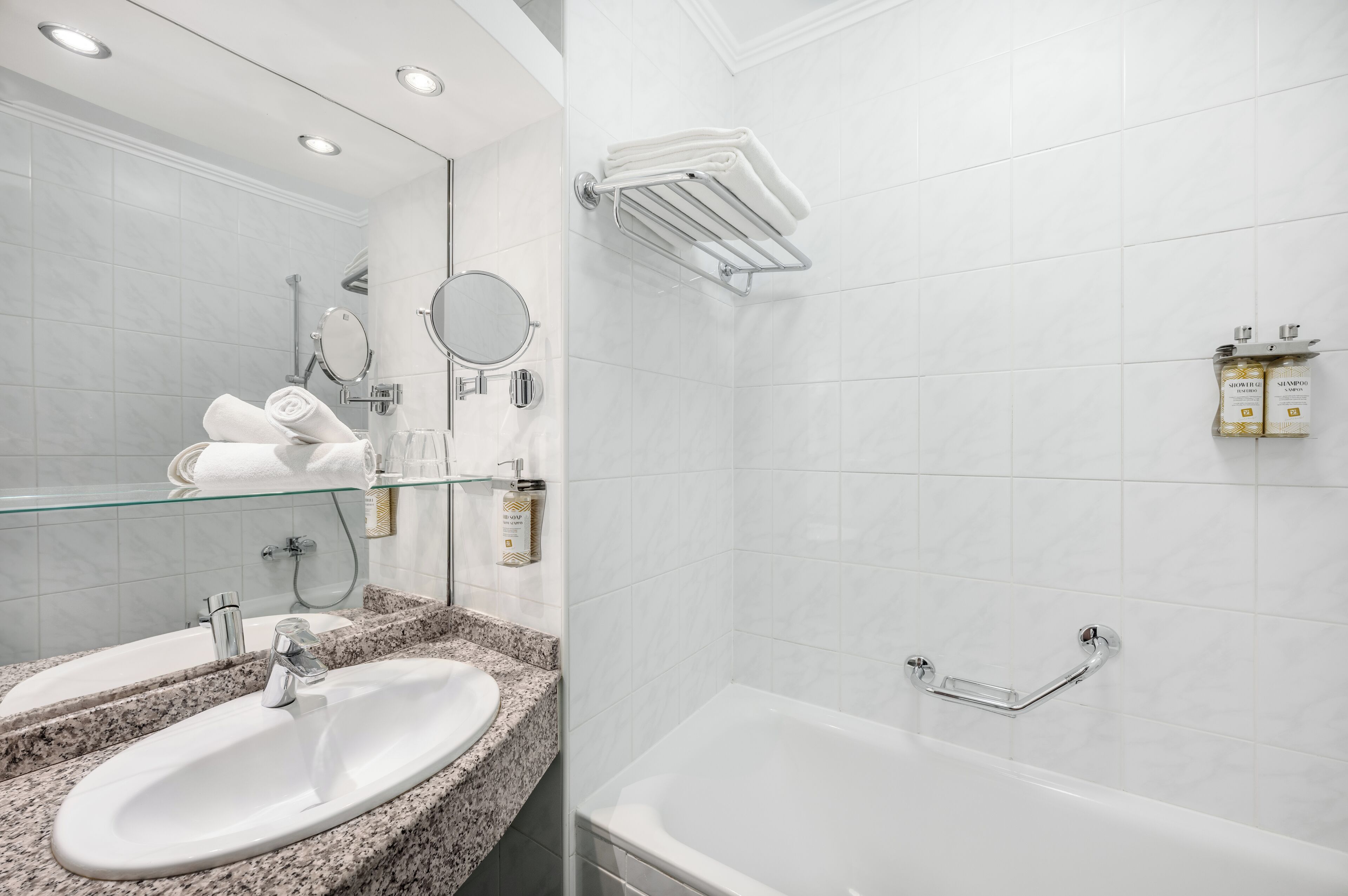 Un baño moderno con lavamanos de encimera de granito, espejo con puerta de cristal, cabezal de ducha de mano y bañera blanca con barra de apoyo cromada.
