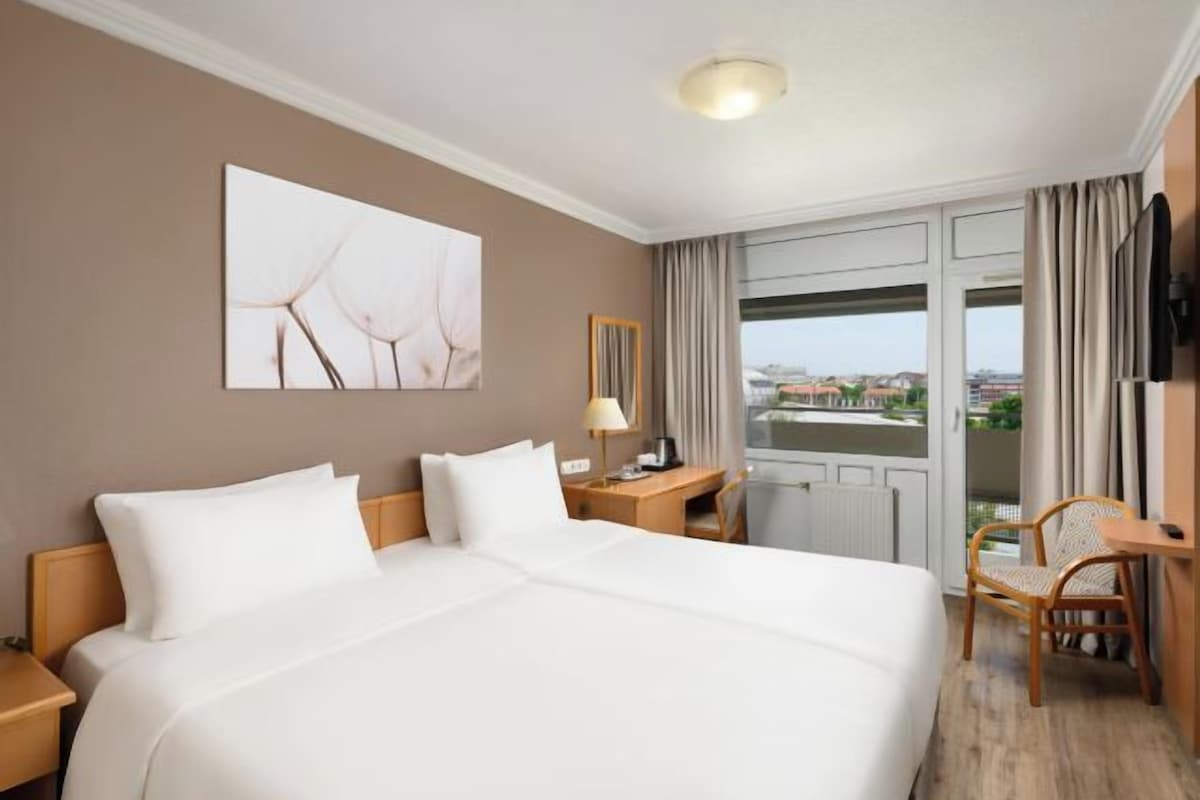 A hotel room with a large bed, a desk, a chair, a window with curtains, and a city view.