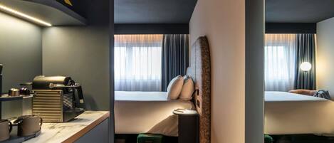 Una habitación de hotel moderna con kitchenette, una cama con cabecero, una mesita de noche y un ventanal con cortinas.