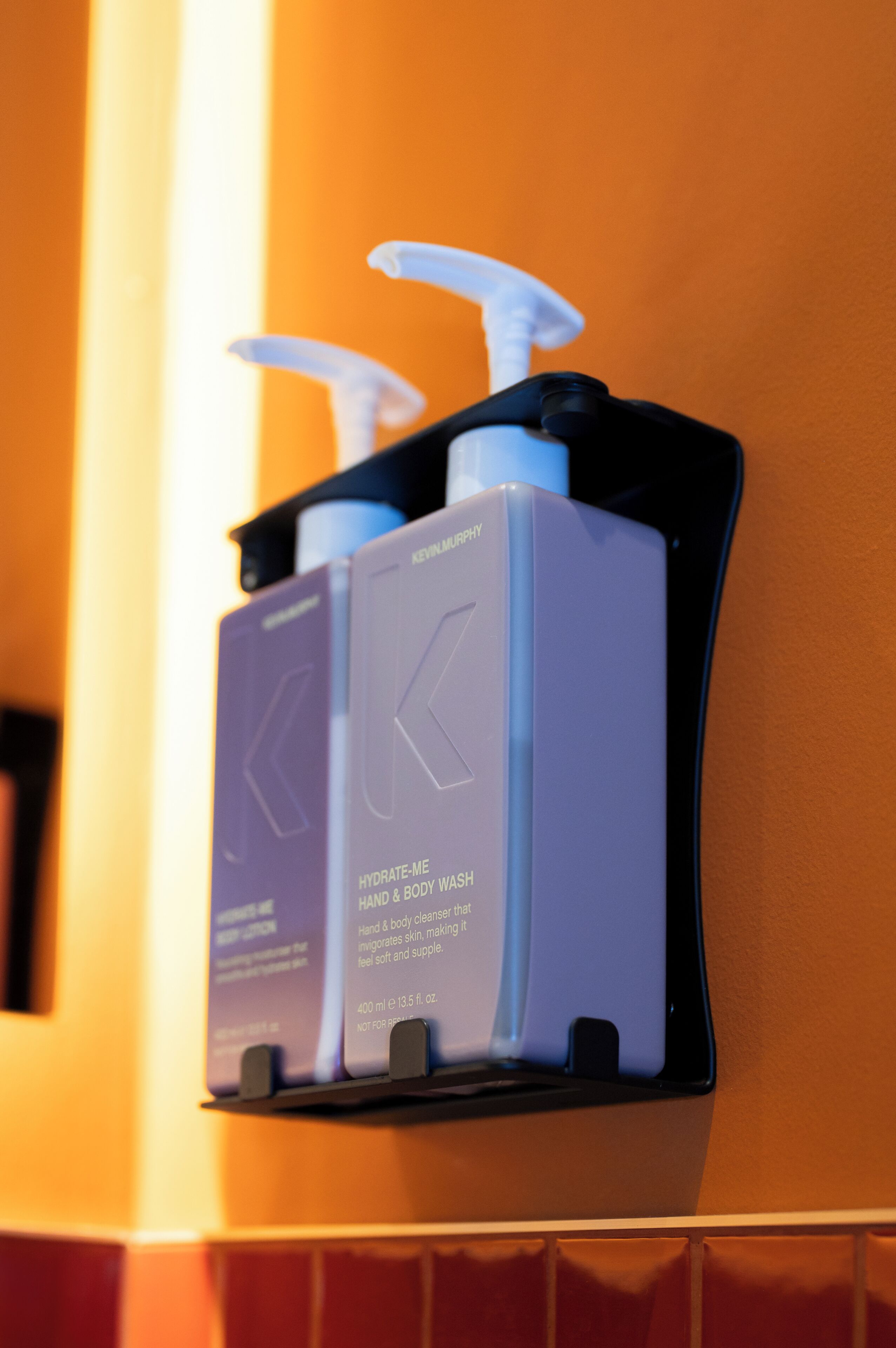 Dispensador montado en la pared con dos botellas etiquetadas “HYDRATE-ME HAND & BODY WASH” de KEEVILMANY.