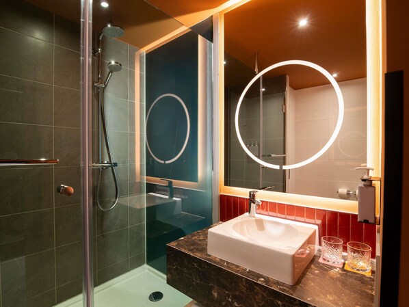 Un baño moderno con ducha acristalada, un espejo grande con luz circular y encimera de mármol con lavamanos.