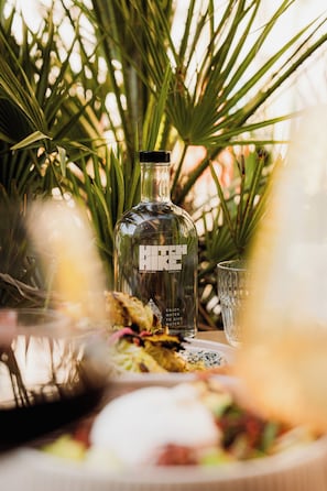 Een fles Hike Coastline gin, een glas en een bord eten op een tafel, met palmbladeren op de achtergrond.