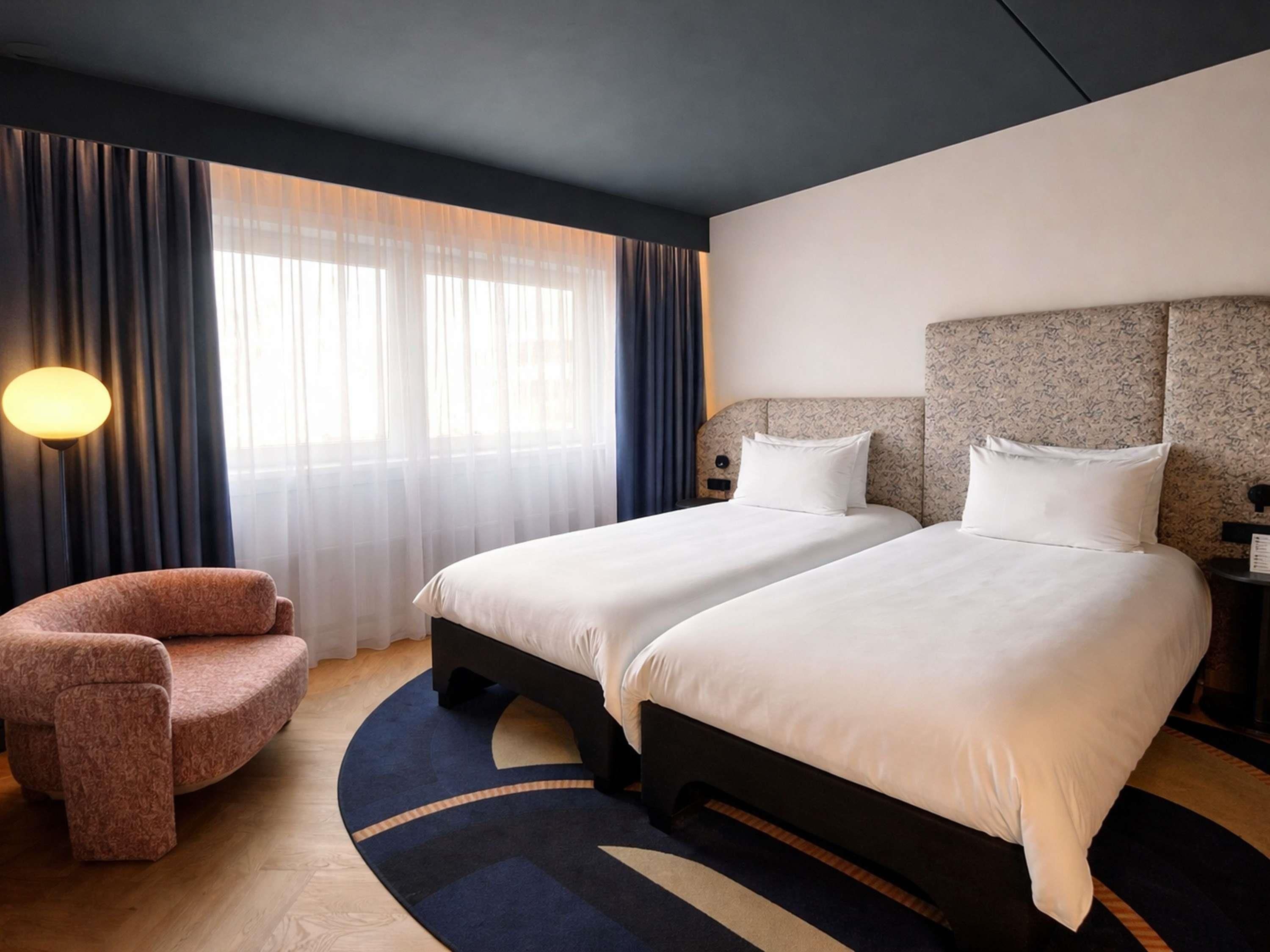 A hotel room with two beds, a chair, a lamp, and a view of the outdoors.