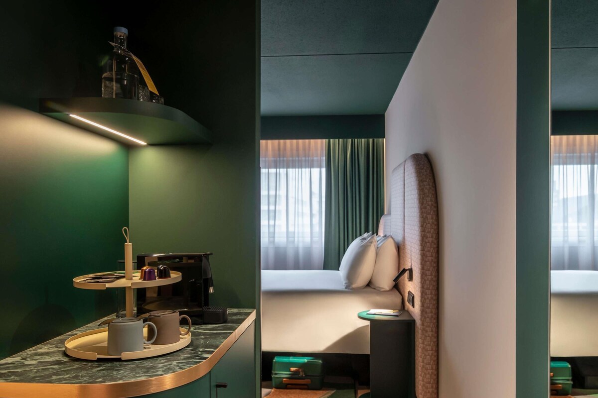 Une chambre d’hôtel avec un bureau incurvé, un lit avec des oreillers, une fenêtre avec des rideaux et un mur vert.