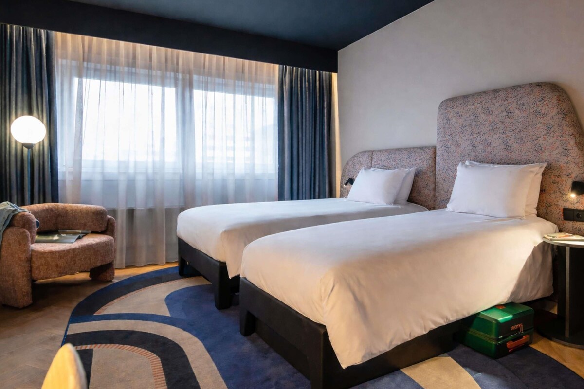 Une chambre d’hôtel avec deux lits, un canapé, une lampe et une vue sur la ville grâce à de grandes fenêtres.