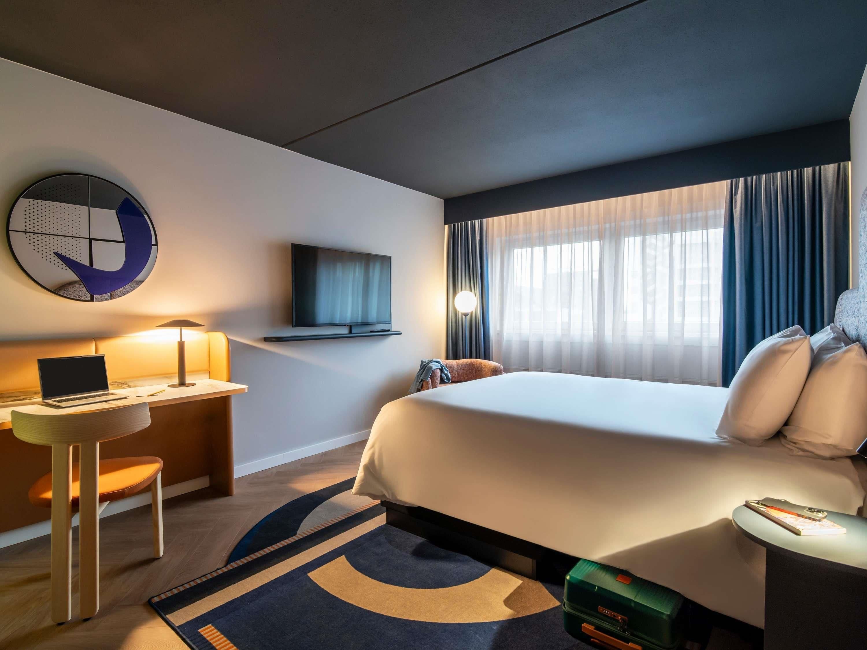 A hotel room with a bed, a desk with a laptop, a TV mounted on the wall, a window with curtains, and a round wall decoration.