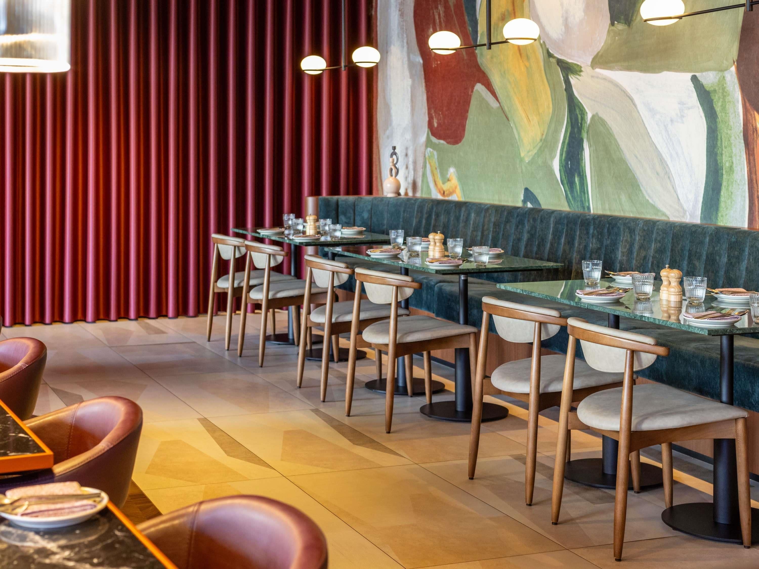 A restaurant interior with a long dining table, cushioned chairs, and a colorful abstract mural on the wall.