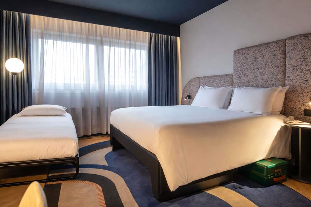 Une chambre d’hôtel avec deux lits, une grande fenêtre avec des rideaux légers, une tête de lit décorée et une petite table avec une lampe.