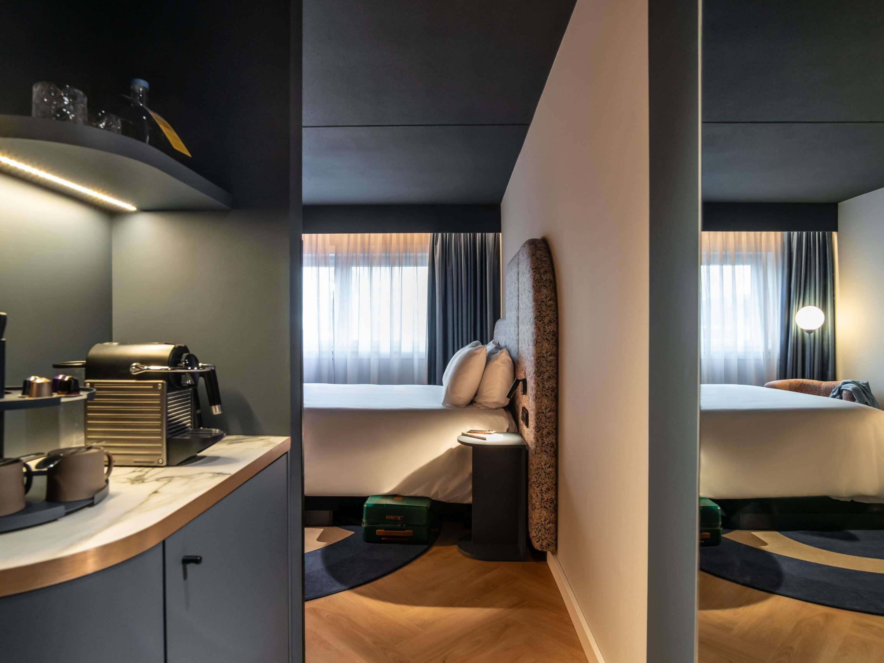 Habitación de hotel con kitchenette, cama con cabecera, mesita de noche, ventana con cortinas y lámpara fijada a la pared.