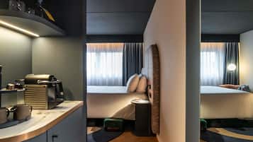 A hotel room with a kitchenette, a bed with a headboard, a bedside table, a window with curtains, and a wall-mounted lamp.