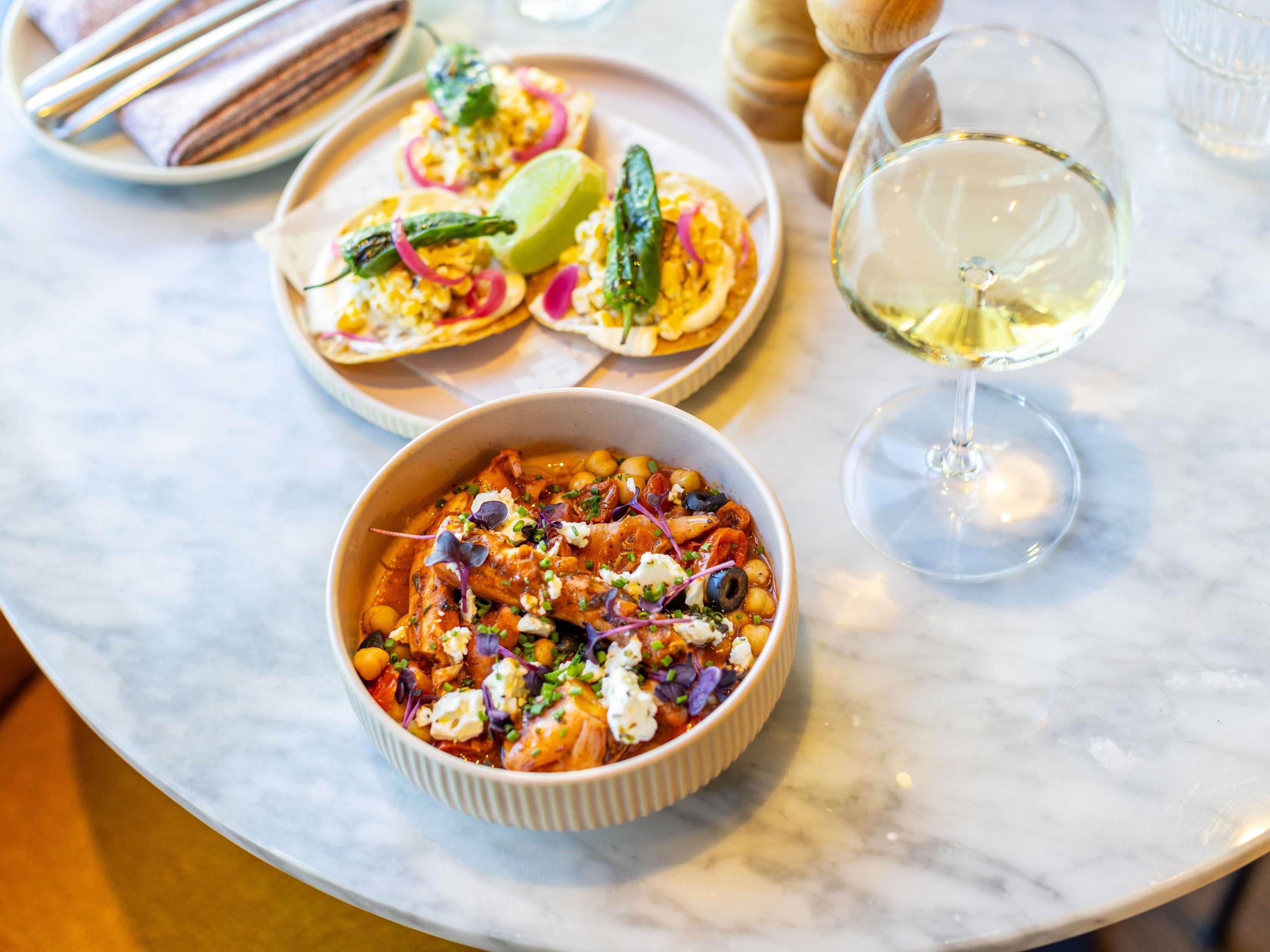 A table with a bowl of food, a plate of tacos, and a glass of white wine.