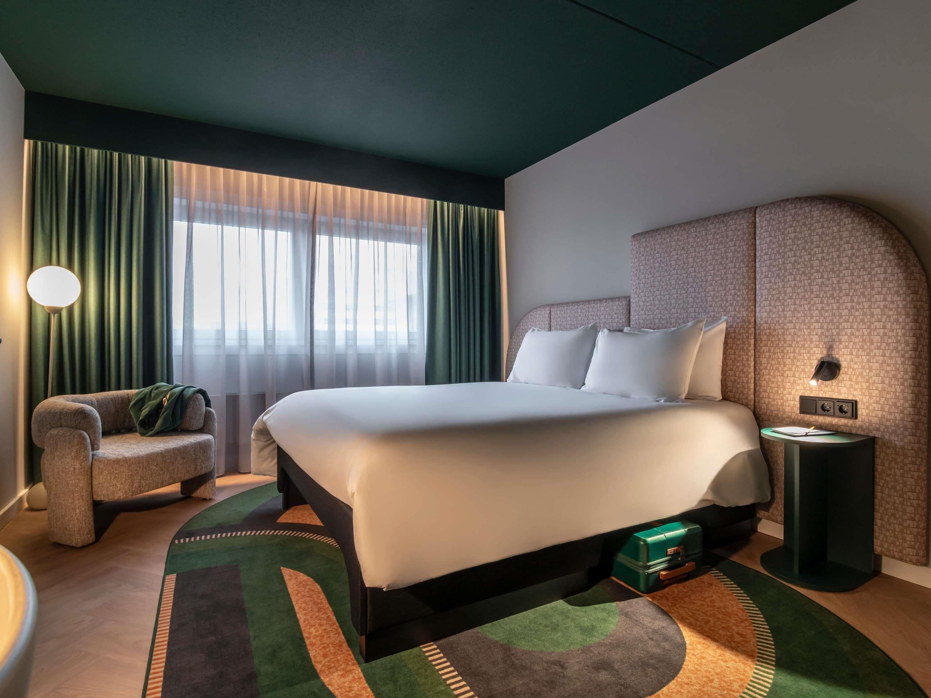 Une chambre d’hôtel moderne avec un grand lit, une table de chevet, une chaise et une vue sur la ville à travers de fines rideaux.