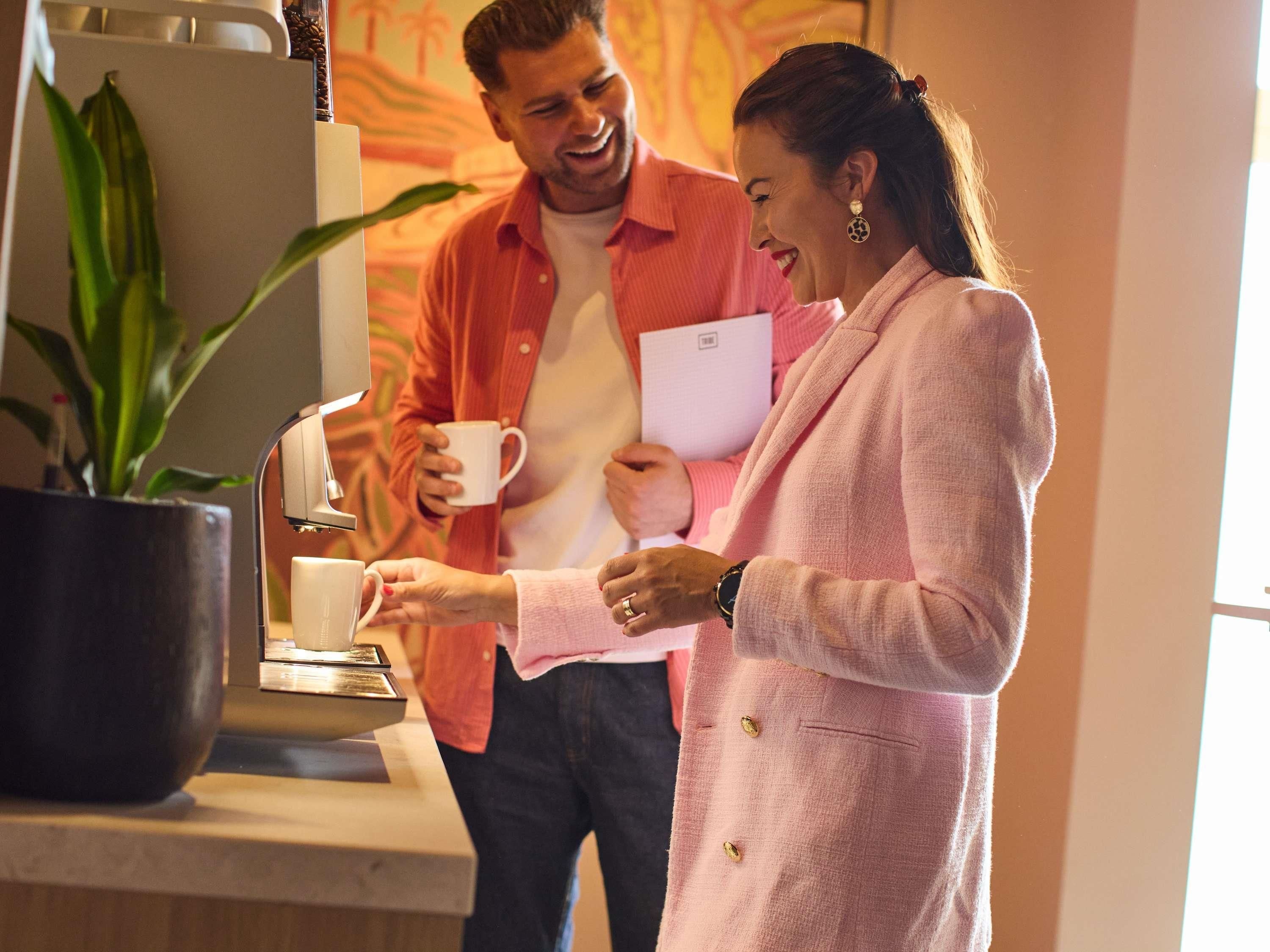 A person in a pink blazer holding a white mug, standing next to a coffee machine with a large potted plant beside it.