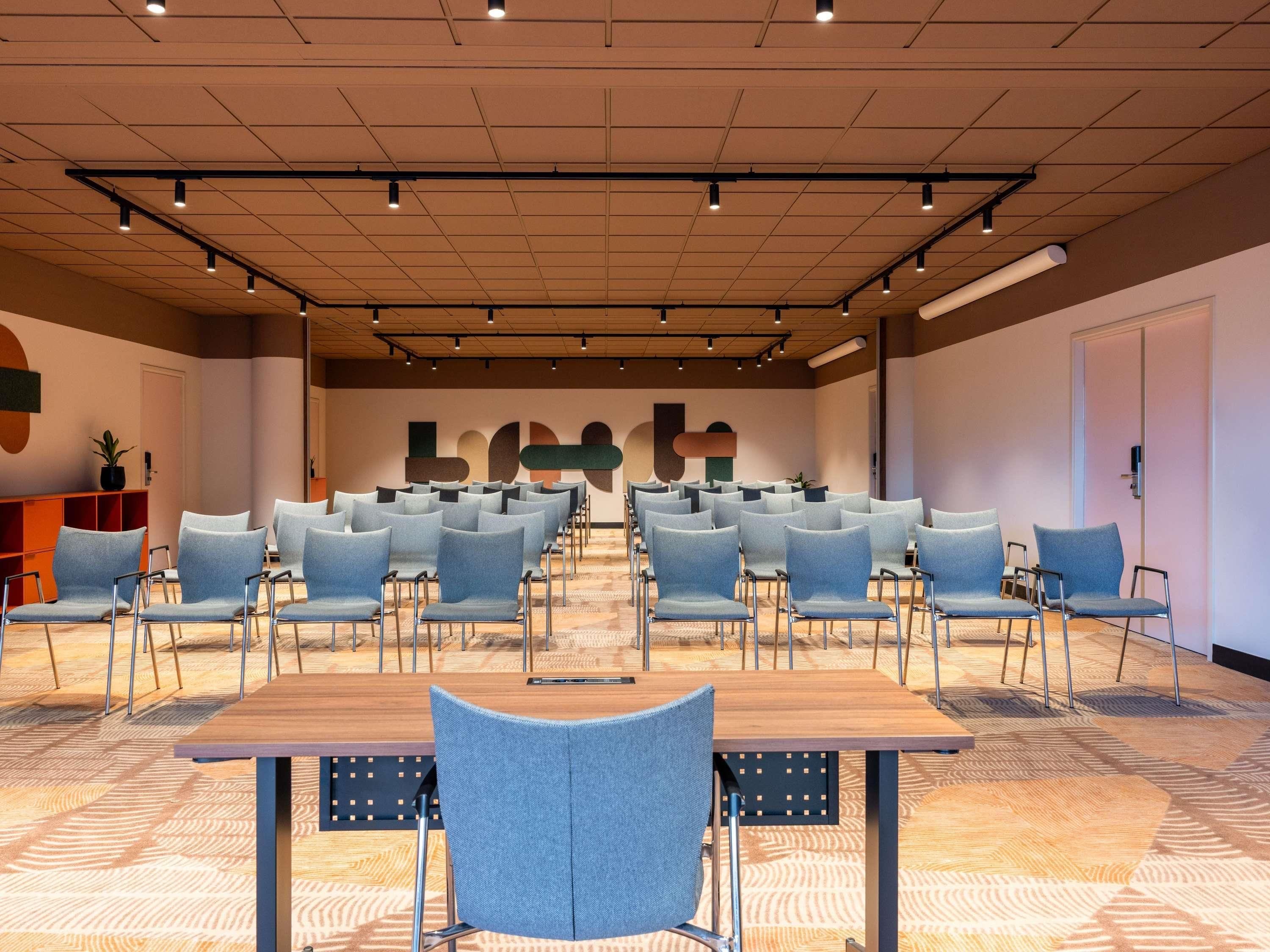 Une salle de conférence avec des rangées de chaises, une table en bois et un tapis à motifs.