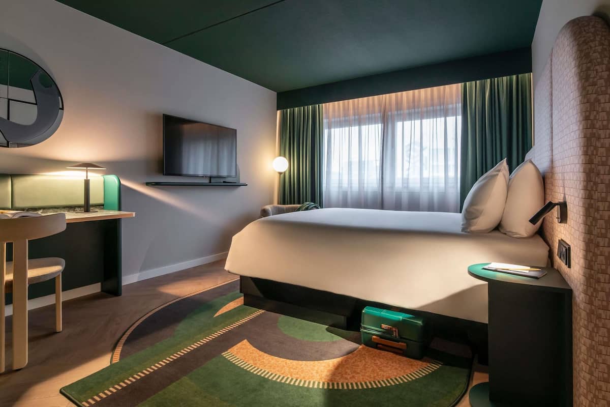 Une chambre d’hôtel avec un lit, un bureau doté d’une lampe, une télévision et une fenêtre avec des rideaux.