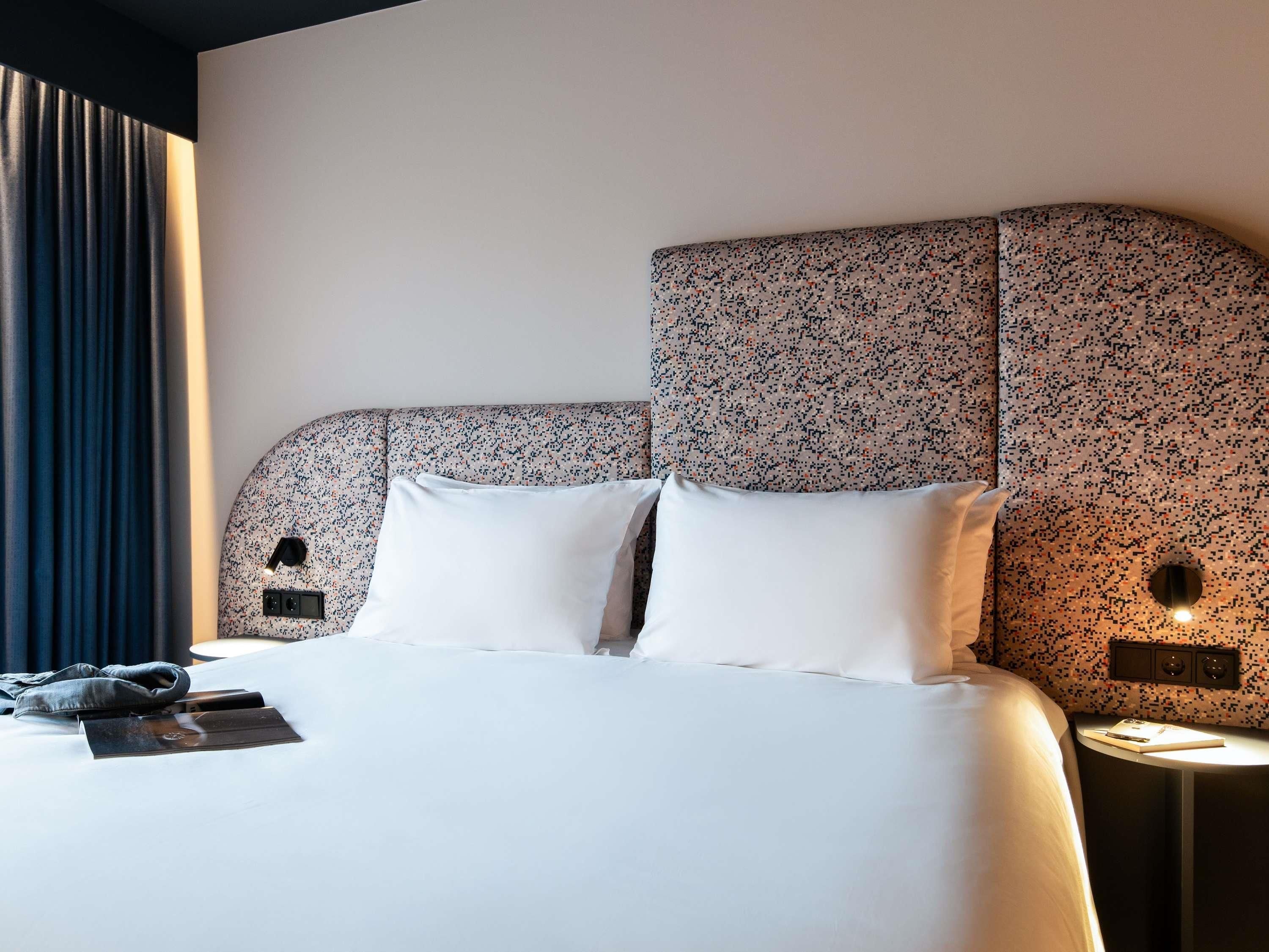 A hotel room with a large bed, a headboard with a floral pattern, a bedside table with a lamp, and a window with curtains.