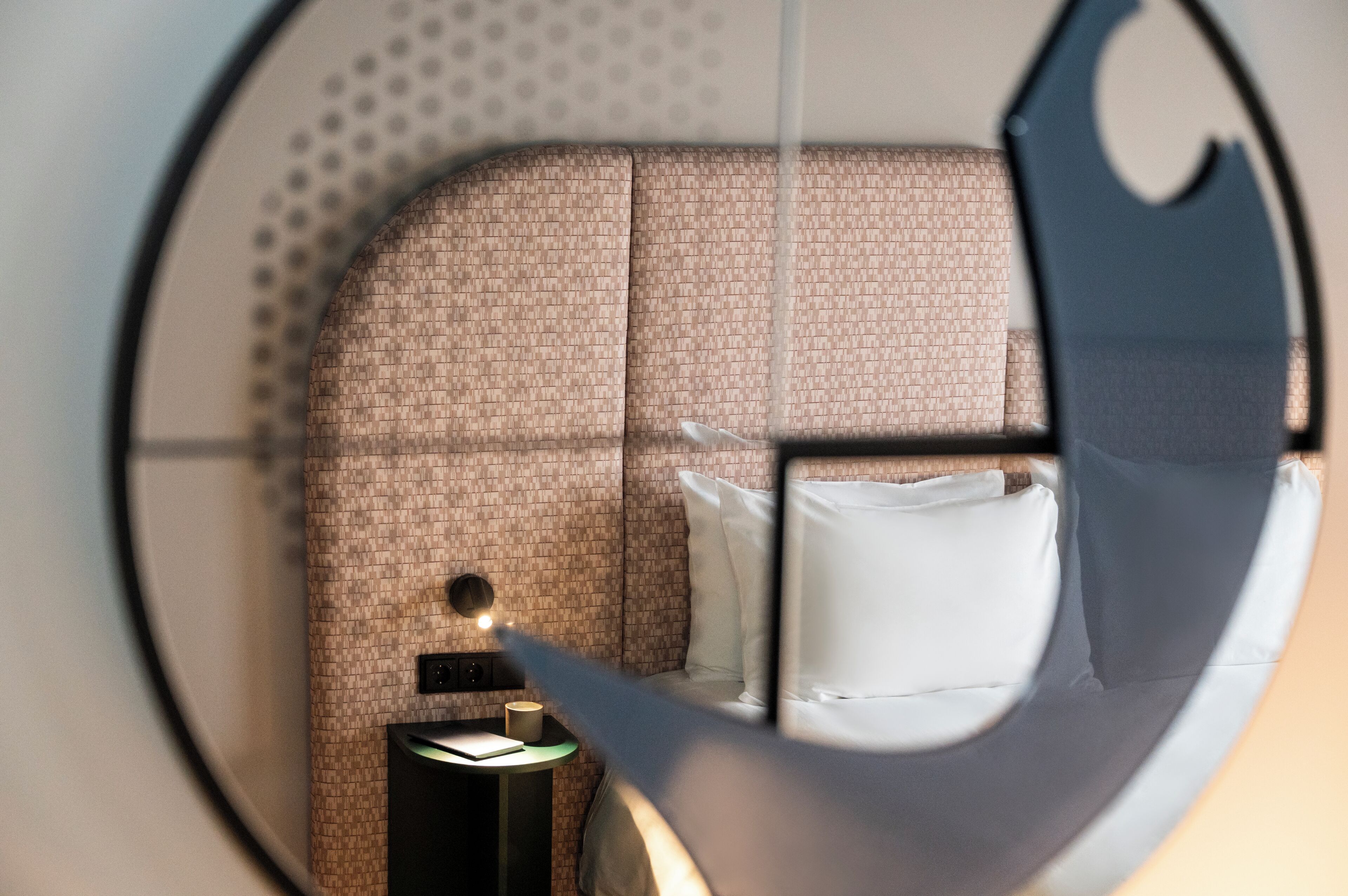 A modern room with a patterned wall, a bed with white bedding, a nightstand with a lamp, and a circular window view.