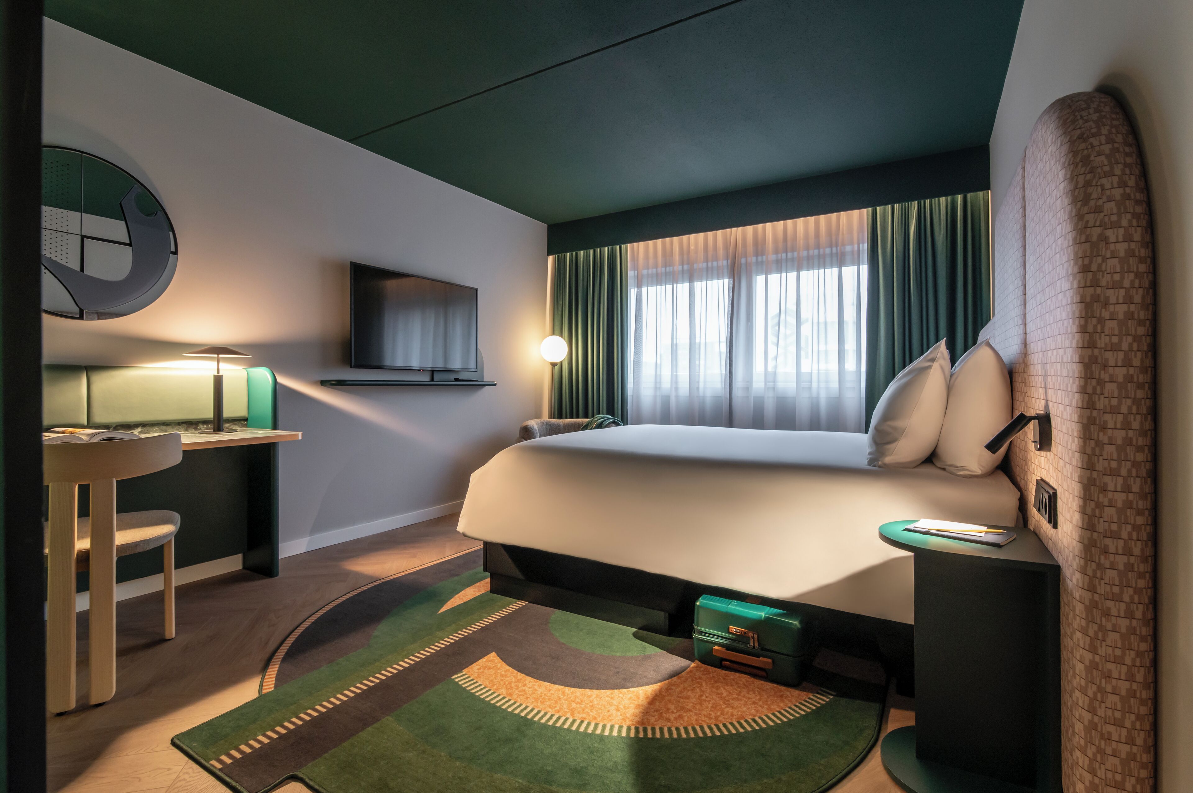 Une chambre d’hôtel moderne avec un grand lit, un bureau avec une lampe, une télévision et une moquette verte.
