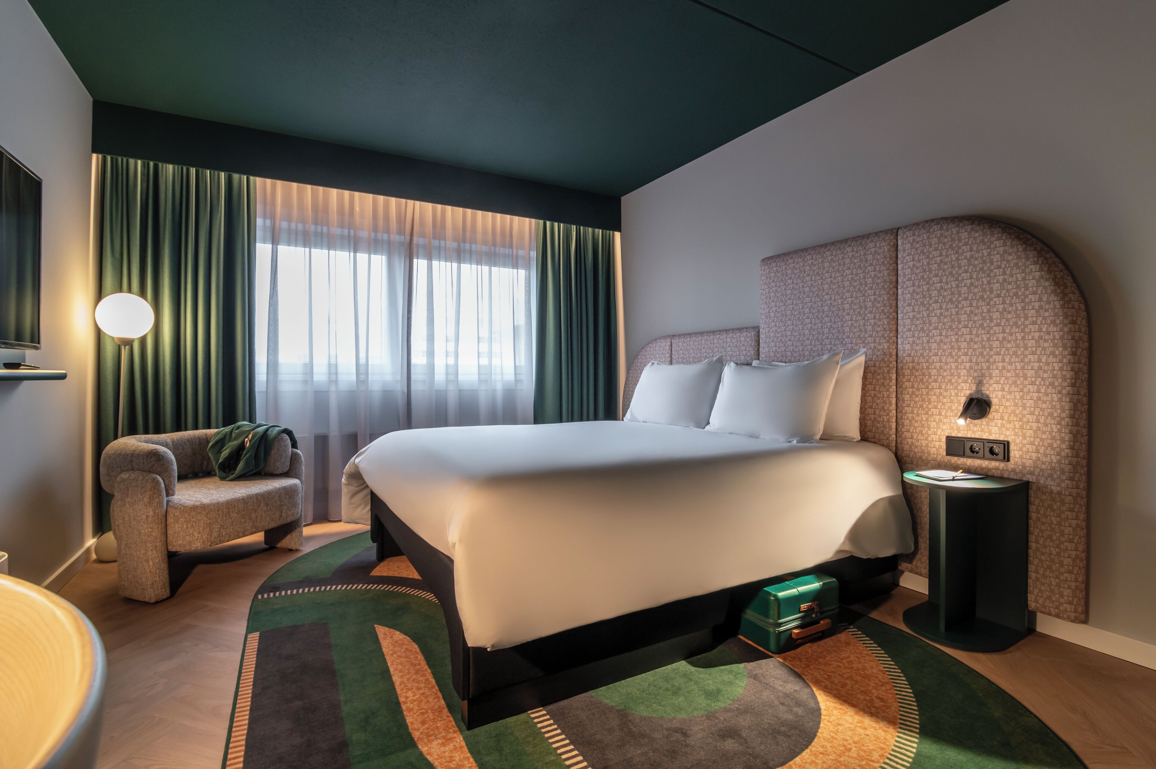 A modern hotel room with a large bed, a sofa, a bedside table, and a lamp.