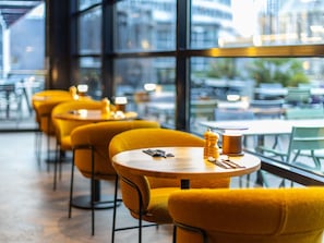 Una cafetería moderna con sillas amarillas y mesas redondas, ventanas amplias y vistas panorámicas de la ciudad.