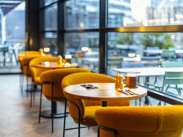 A modern cafe with yellow chairs and round tables, large windows, and a view of the cityscape.