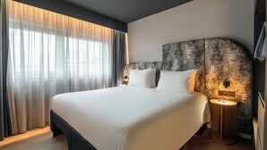 Een moderne hotelkamer met een groot bed, een hoofdeinde met een getextureerde afwerking, een nachtkastje met een lamp en lichte gordijnen voor het raam.