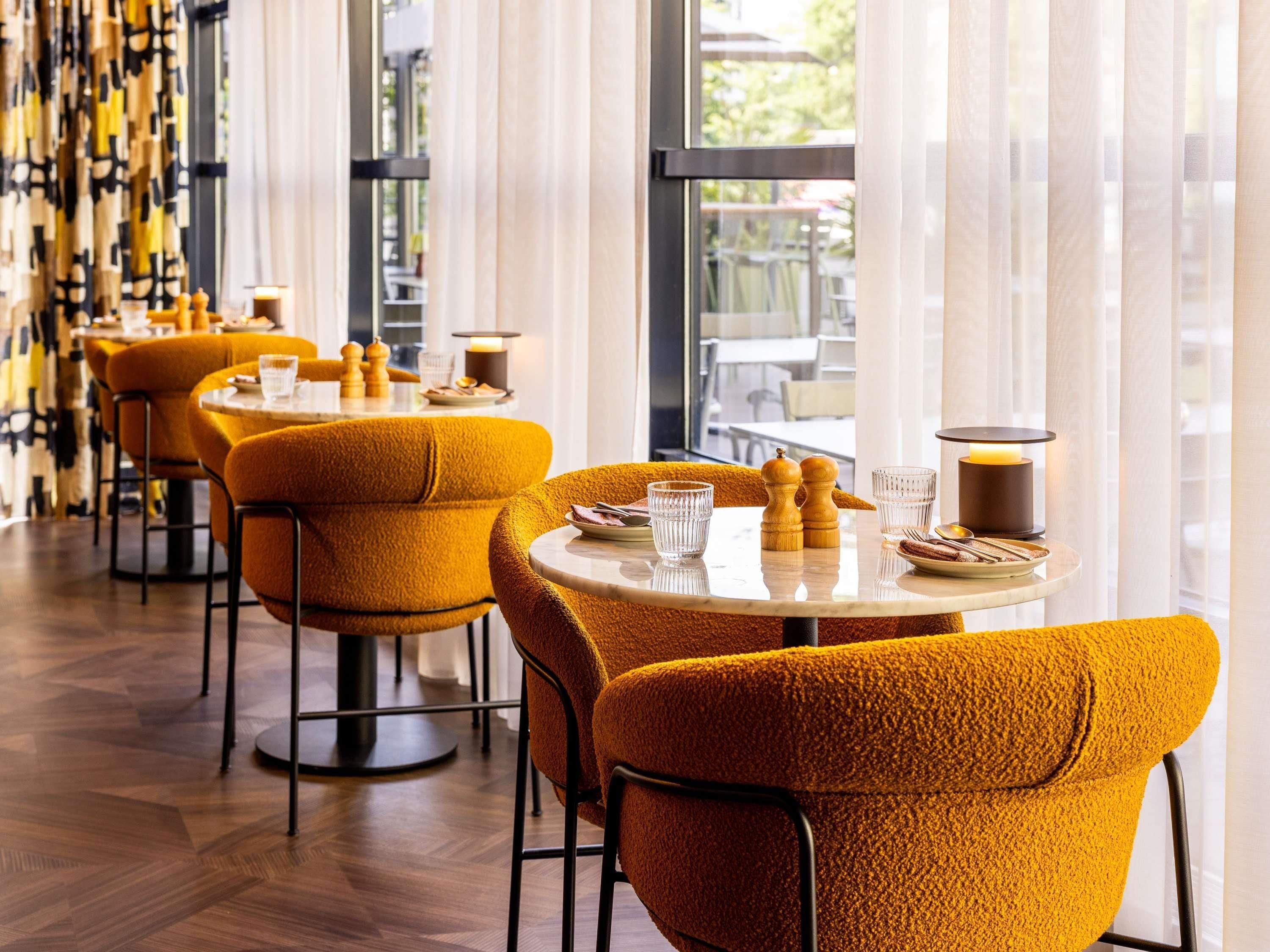 A restaurant with round tables, orange chairs, and a view through large windows.