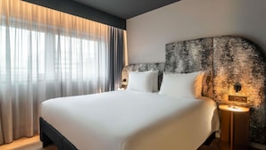 A modern hotel room with a large bed, a headboard with a textured pattern, a bedside table with a lamp, and sheer curtains on the window.