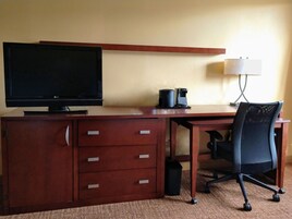 Habitación de hotel con escritorio de madera, televisor, cafetera, lámpara y una silla de oficina negra.