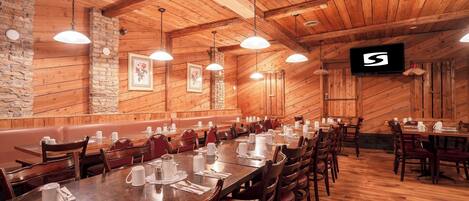 A restaurant with wooden walls and ceiling, long tables set with white tableware, and hanging pendant lights.