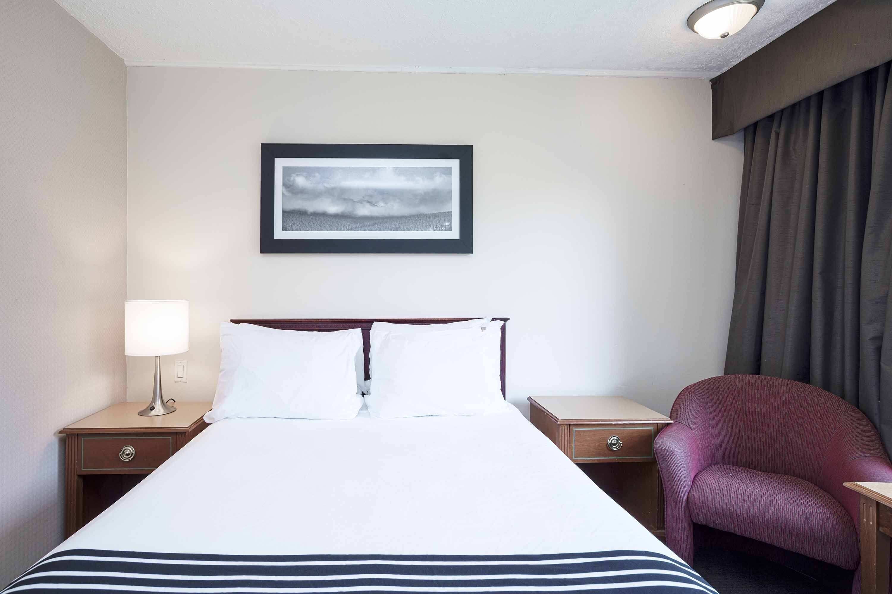 A hotel room with a bed, a nightstand with a lamp, a chair, and a framed picture on the wall.