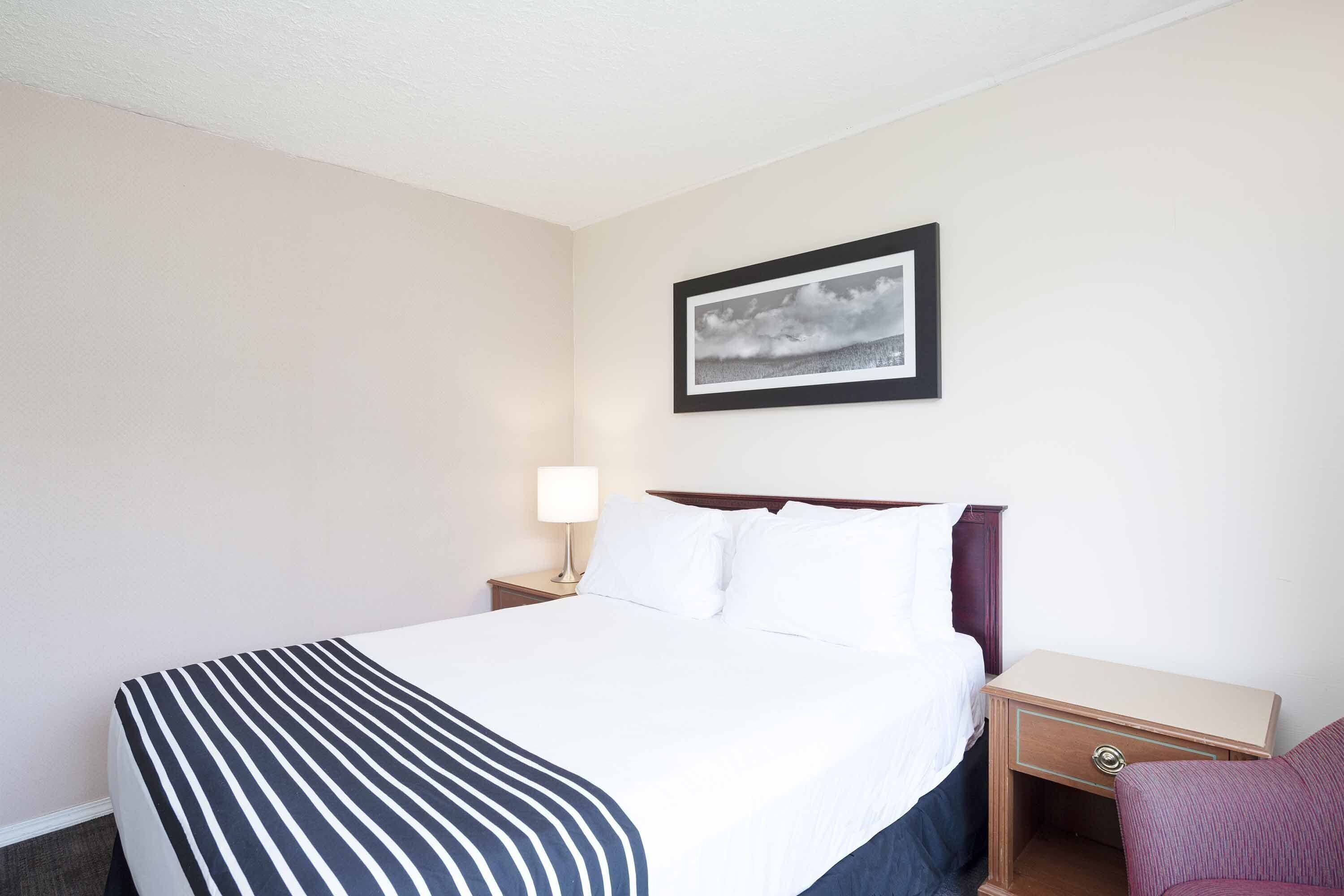 A hotel room with a bed, a nightstand with a lamp, a framed picture on the wall, and a chair.