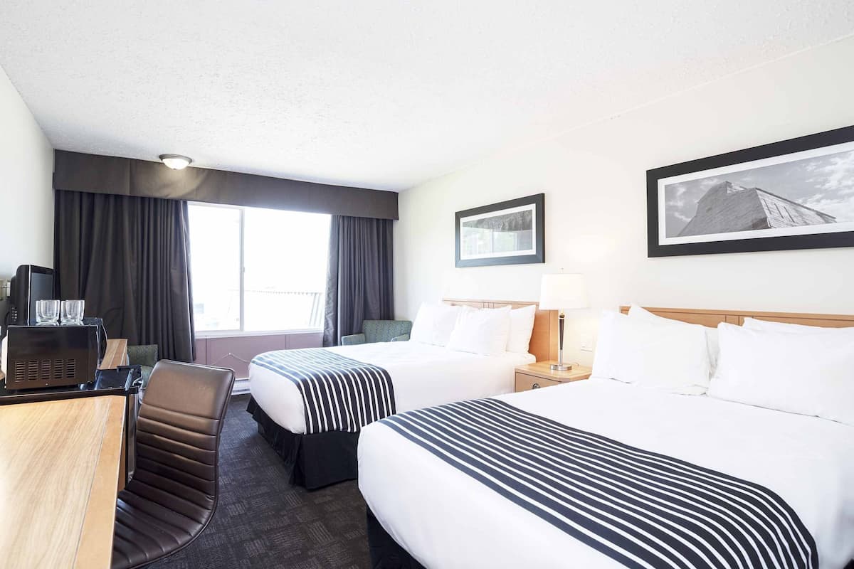 A hotel room with two beds, a desk with a computer, a chair, a window with curtains, and a framed picture on the wall.