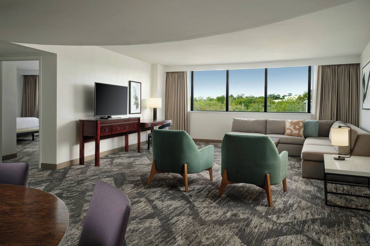 Habitación de hotel con sofá, dos sillones, televisor, escritorio con lámpara y ventana grande con vistas a zonas verdes.