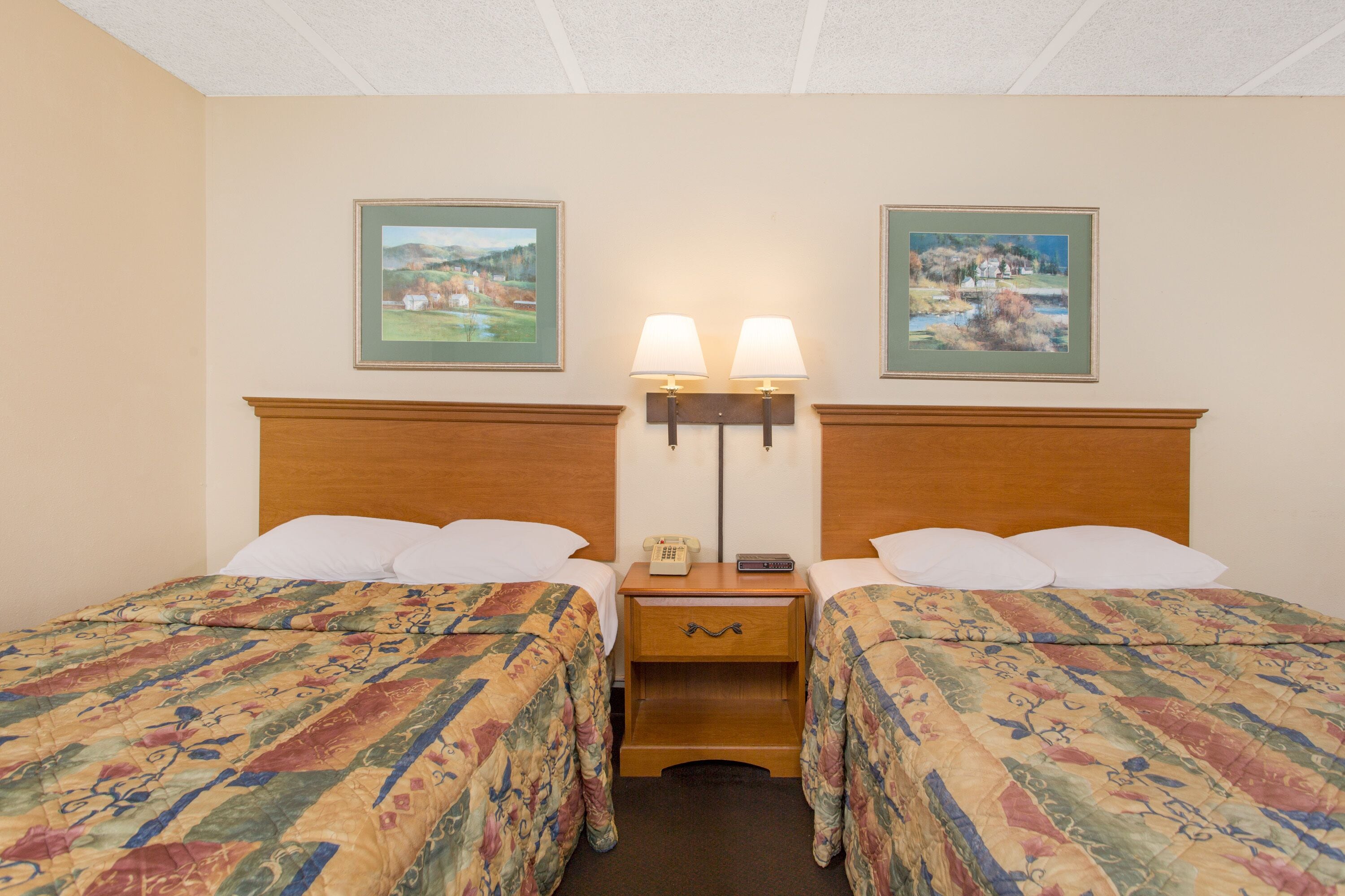 A hotel room with two beds, each with a floral-patterned bedspread, a wooden headboard, a nightstand with a lamp, and two framed pictures on the wall.