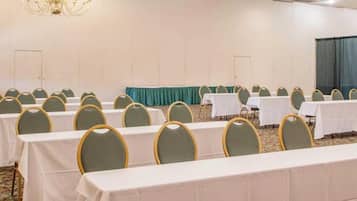 Un salón de conferencias con filas de mesas y sillas, una araña y una cortina verde.