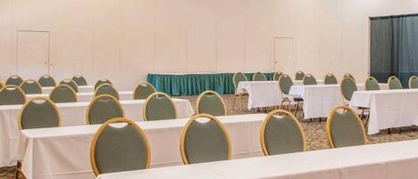 Un salón de conferencias con filas de mesas y sillas, una araña y una cortina verde.