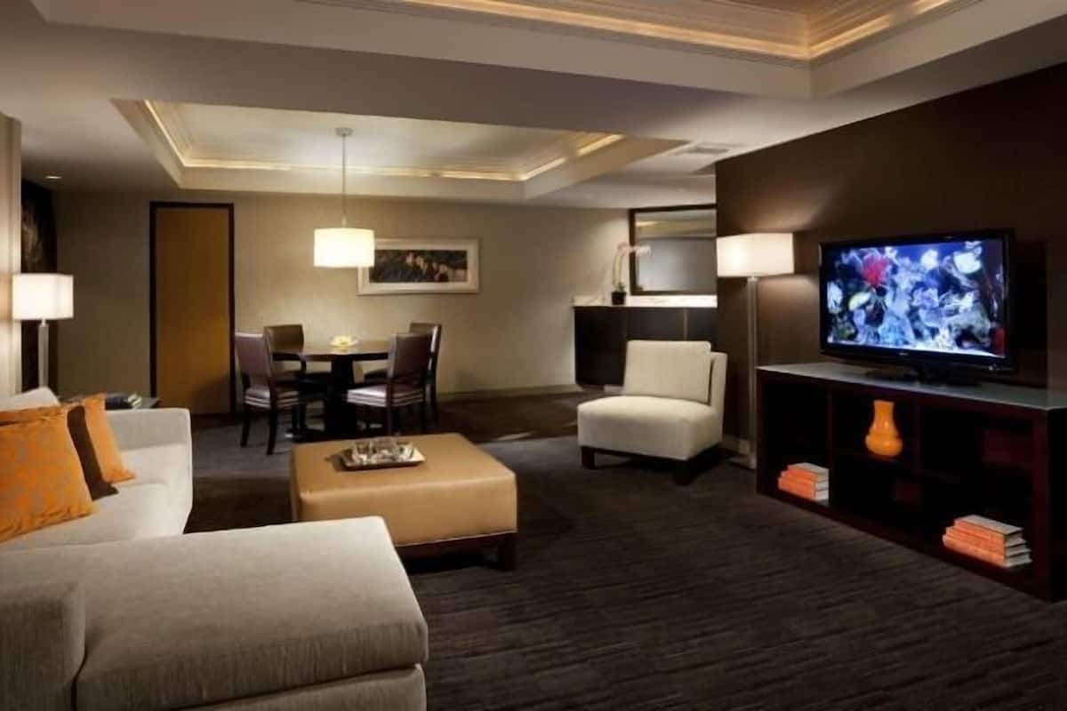 A hotel room with a sofa, ottoman, dining table, chairs, a TV, and a bookshelf.