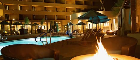 A pool area with lounge chairs, umbrellas, and a fire pit.